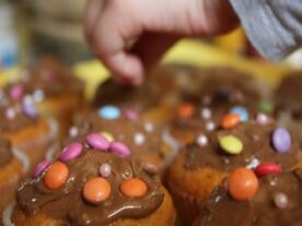 Aszalt meggyes muffin: Kézzel díszített, csokoládéval bevonatott muffinok színes cukorkákkal a tetején, fókuszban egy éppen díszített darab.