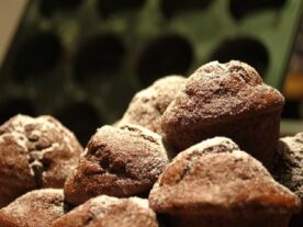 Porcukorral meghintett sötétbarna banános-étcsokis muffinok láthatók a képen, előtérben éles fókusszal és homályos háttérrel, melyben egy muffinforma sejlik fel.