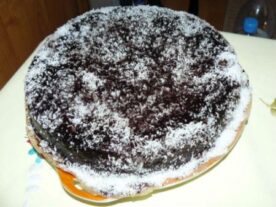 Bounty torta: Kör alakú, sötét csokoládéval bevont torta, bőségesen megszórva fehér kókuszreszelékkel a tetején, tálalva egy színes tortatányéron.