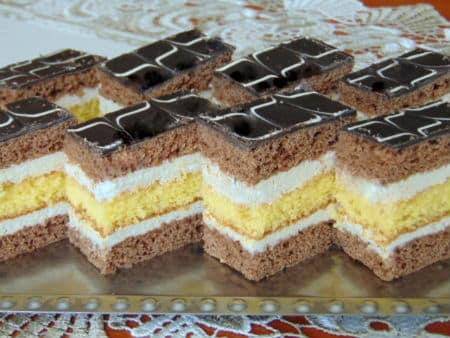 Többrétegű "Vendégváró szelet" sütemény, váltakozó barna kakaós és sárga piskóta rétegekkel, fehér krémtöltelékkel, tetején sima csokoládémázzal, darabonként szabályos négyzet alakúra vágva.