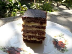 Stollwerck szelet: Egy réteges sütemény szelet, világos és sötét barna rétegekkel váltakozva, porcukorral meghintve, tálalva egy virágmintás porcelántányéron, természetes fényben fotózva.