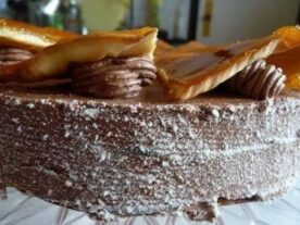 Egy Dobos torta részletgazdag képe, ami sötét csokoládé vajkrémmel rétegezett világos színű piskóta, a tetején karamellel díszítve, ami szép kontrasztot ad a csokoládé színével. A torta oldalát apró csokoládé morzsa borítja, és a szeletek láthatóan egyenletesen vannak elosztva.
