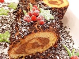 Karácsonyi fatörzs sütemény porcukorral megszórva, csokoládé bevonattal és cukormázzal készült piros bogyókkal, zöld levelekkel díszítve, ünnepi tálaláson.