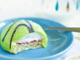 Egy szelet svéd Prinsesstårta zöld marcipán bevonattal, málnalekvárral és tejszínhabbal rétegezve, díszítve csokoládécsíkokkal, tálalva egy világoskék tányéron egy világos színű asztali abrosszal a háttérben.