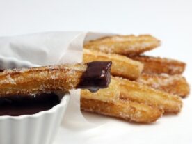 Frissen sült churrosok cukorral meghintve egy fehér papírral bélelt tányéron, mellettük egy kis tál sötét csokoládé mártás.