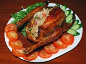 Jól megsült, aranybarna Bugaci töltött csirke tálalva friss zöldségekkel, paradicsommal és uborkával egy fehér tányéron, töltelékkel a tál közepén.